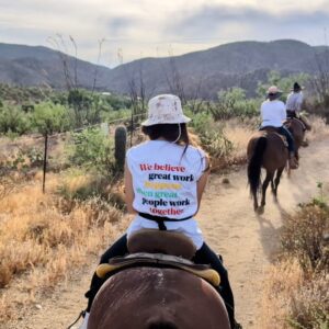 Woman on a horse from behind. Her shirt reads, "we believe great works happens when people work together."