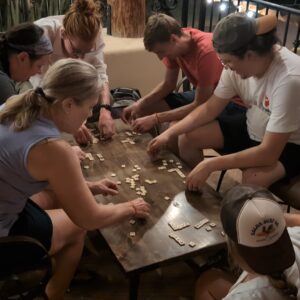 6 people crowded around a small coffee table, intently playing Bananagrams